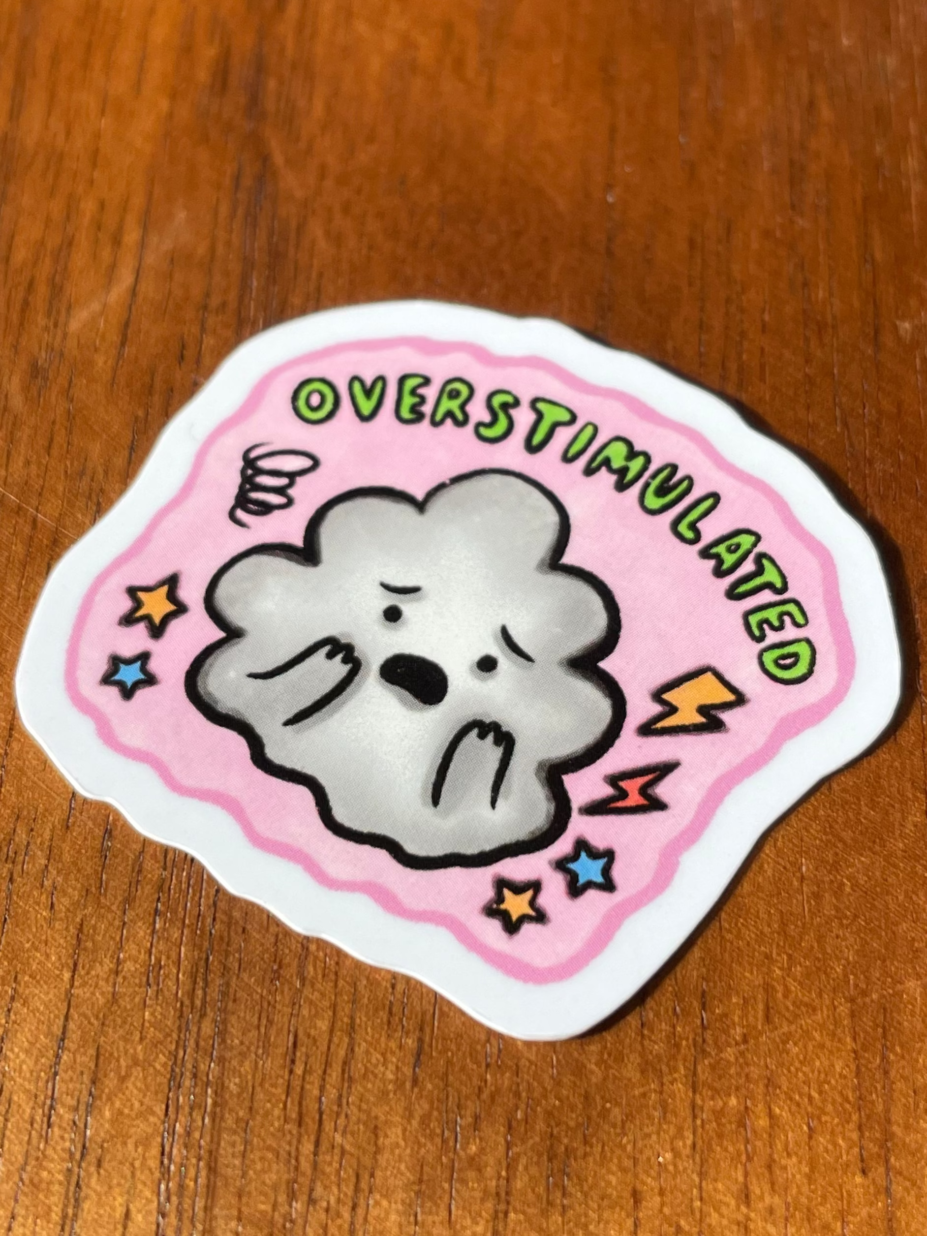 A die-cut sticker featuring a cartoon gray cloud with a sad face on a pink background, with the text 'OVERSTIMULATED' and small lightning bolts and stars, placed on a wooden surface.