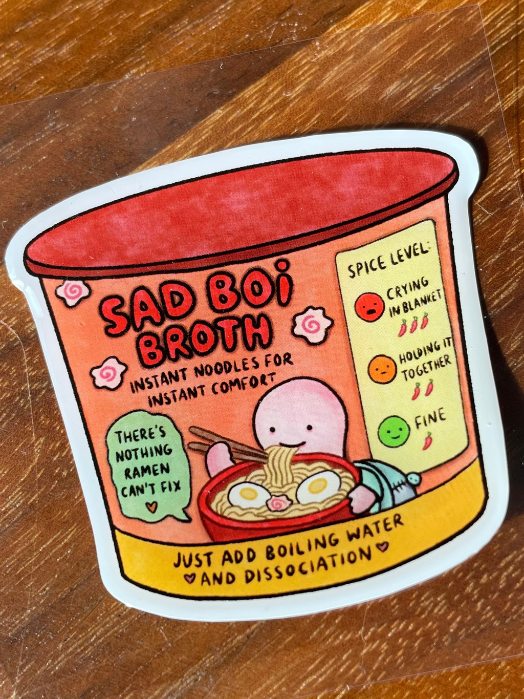A die-cut sticker featuring a cartoon cup of ramen with the text 'BAD BOI BROTH' and humorous labels, placed on a clear sheet over a wooden tabletop.