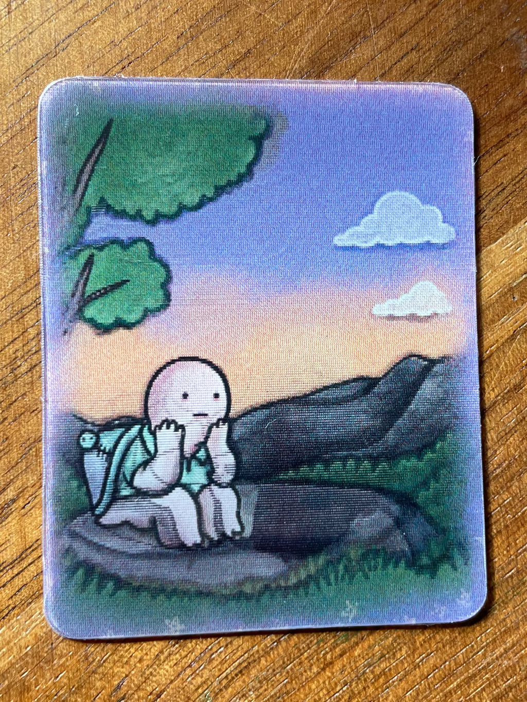 A square illustrated card or sticker with rounded corners, featuring a cartoon figure sitting on a rock in a landscape with trees, mountains, and clouds. The card is placed on a wooden surface.