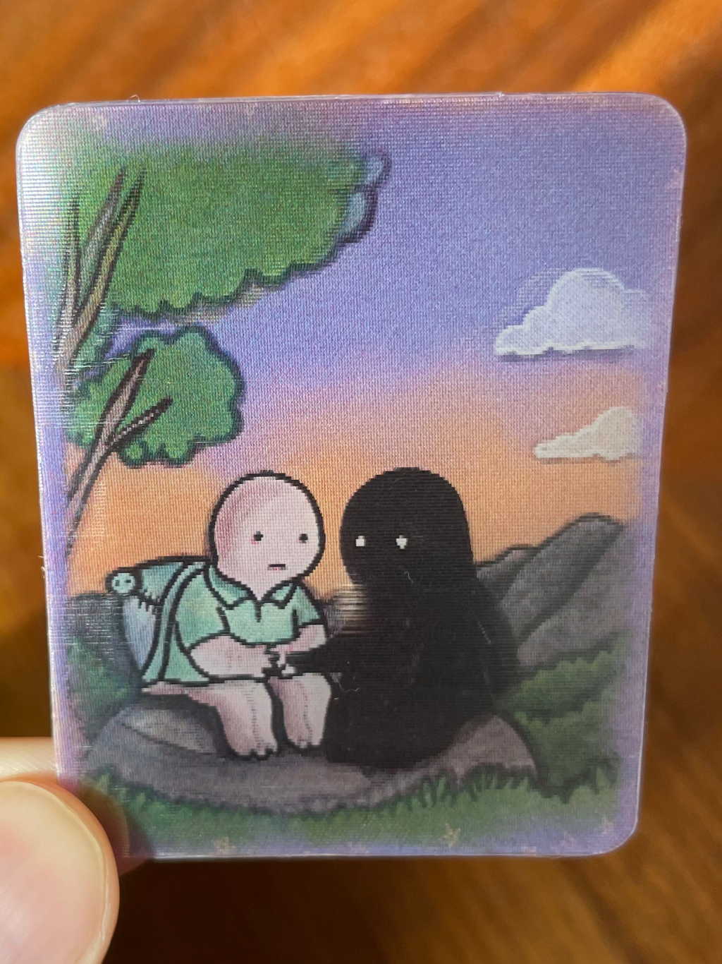 A hand holds a small rectangular illustrated card or coaster featuring a stylized scene of a pale figure and a black shadow-like figure sitting outdoors under a purple sky. The background is a wooden table.